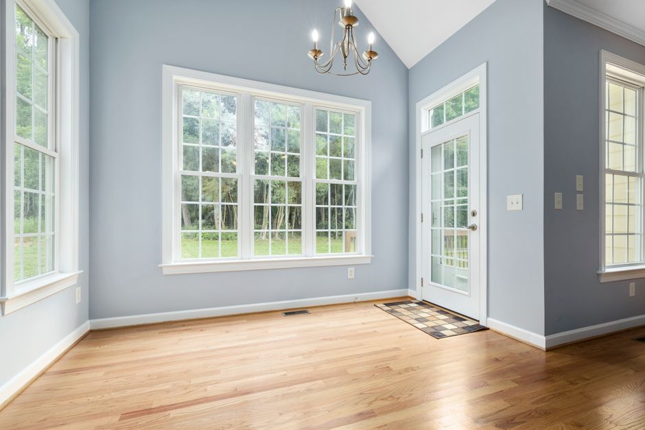 Sunlit empty room with wooden floor, chandelier, and French windows leading outside.